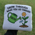 Sweatshirt with a graphic of a watering can labeled 'Faith' and a flower, with text 'Grow Through What You Go Through', held against a grass background.custom-couple-shirt