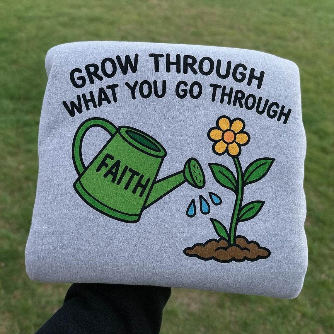 Sweatshirt with a graphic of a watering can labeled 'Faith' and a flower, with text 'Grow Through What You Go Through', held against a grass background.custom-couple-shirt