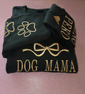 Folded dark green sweatshirt with gold paw prints and 'DOG MAMA' text on a pink surface.custom-couple-shirt