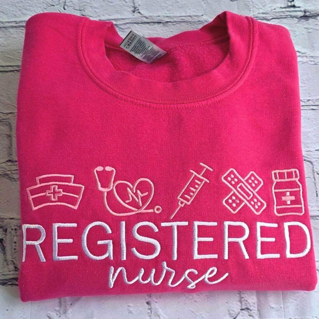 Folded pink sweatshirt with 'Registered Nurse' text and icons on a wooden surface.nurse-embroidered-sweatshirt