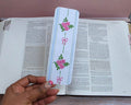 Hand holding a decorative bookmark with floral designs over an open book.coquette-floral-bookmarks