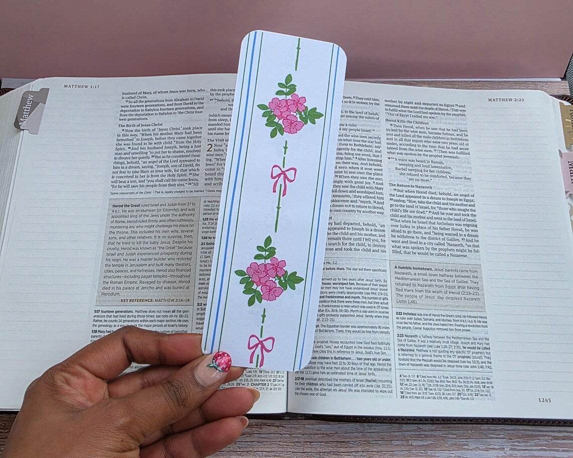 Hand holding a decorative bookmark with floral designs over an open book.coquette-floral-bookmarks