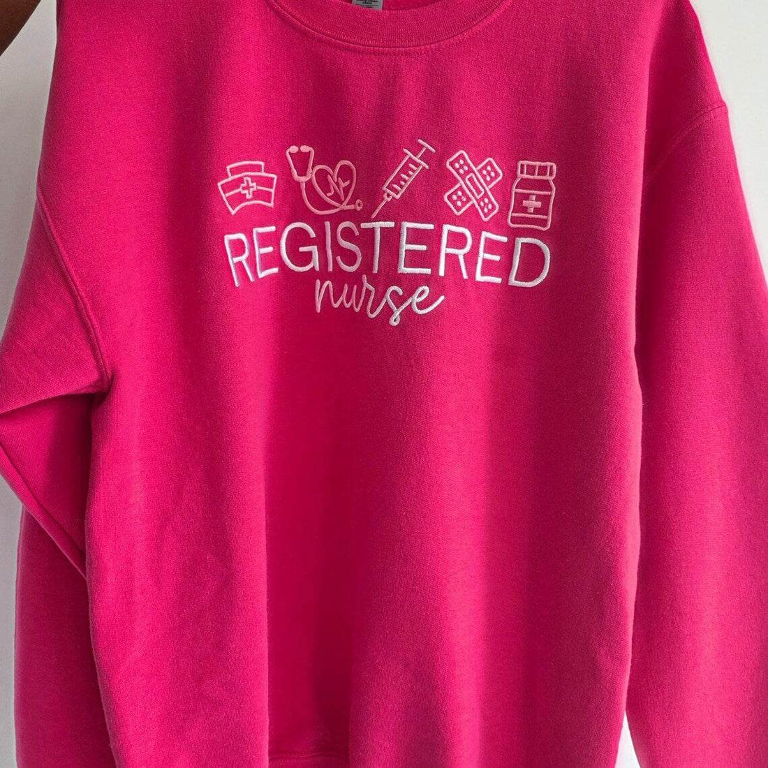 Folded pink sweatshirt with 'Registered Nurse' text and icons on a wooden surface.nurse-embroidered-sweatshirt