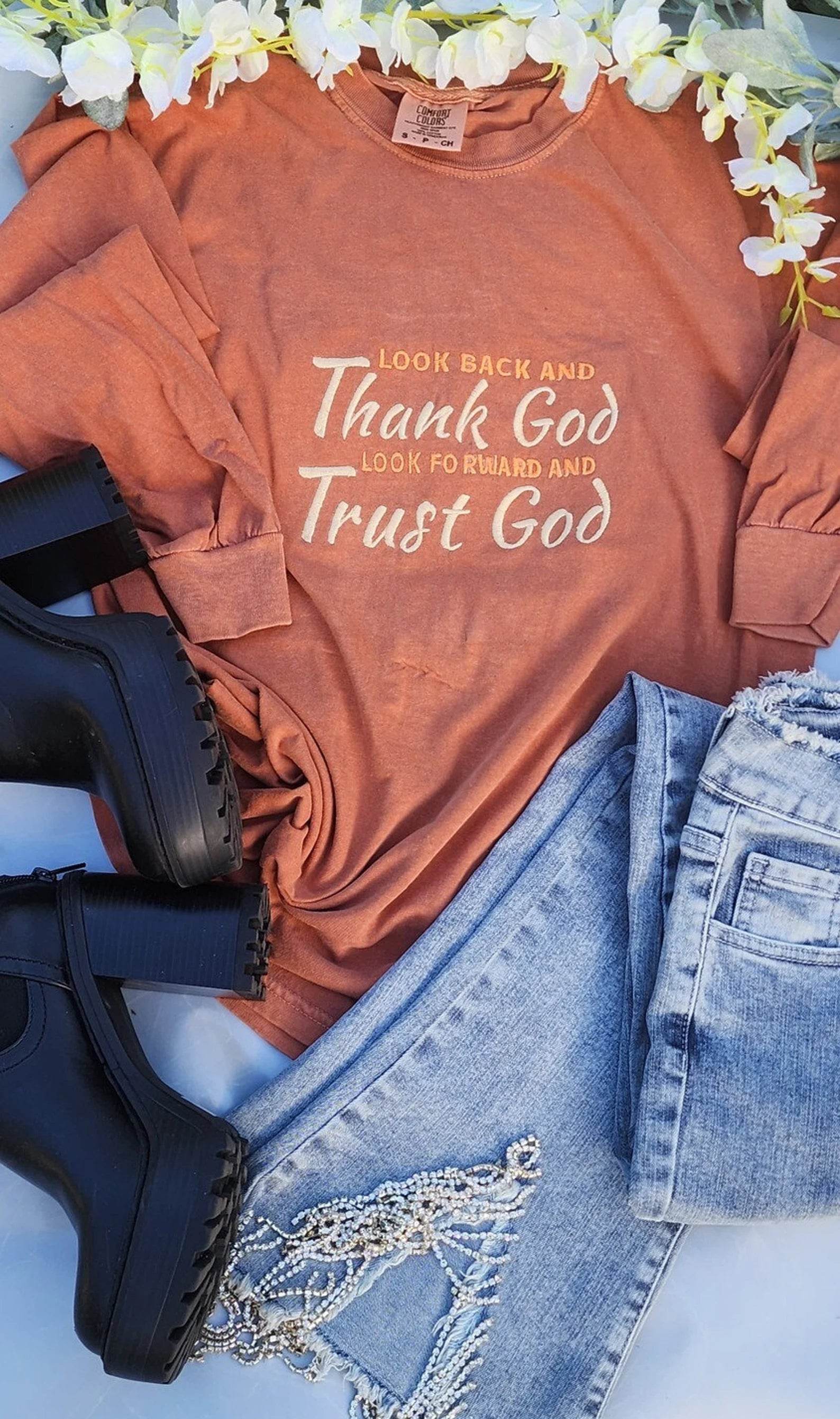 An inspirational t-shirt in brown color with the text 'Thank God, Trust God' printed on the front, displayed alongside denim jeans and black boots, set against a floral backdrop.