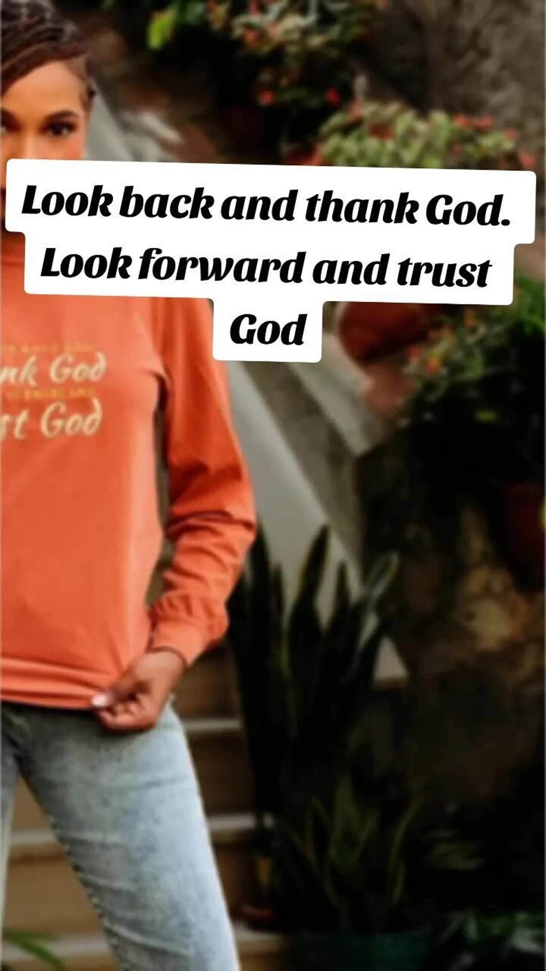 An inspirational t-shirt in brown color with the text 'Thank God, Trust God' printed on the front, displayed alongside denim jeans and black boots, set against a floral backdrop.