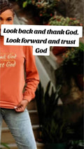An inspirational t-shirt in brown color with the text 'Thank God, Trust God' printed on the front, displayed alongside denim jeans and black boots, set against a floral backdrop.