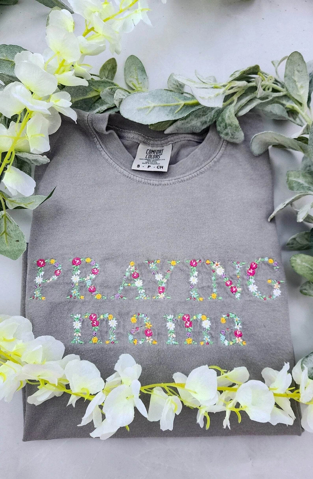 A long sleeve gray t-shirt with the text 'PRAYING MAMA' embroidered in colorful letters, displayed flat with a floral arrangement around it.praying-mama-shirt