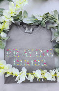 A long sleeve gray t-shirt with the text 'PRAYING MAMA' embroidered in colorful letters, displayed flat with a floral arrangement around it.praying-mama-shirt
