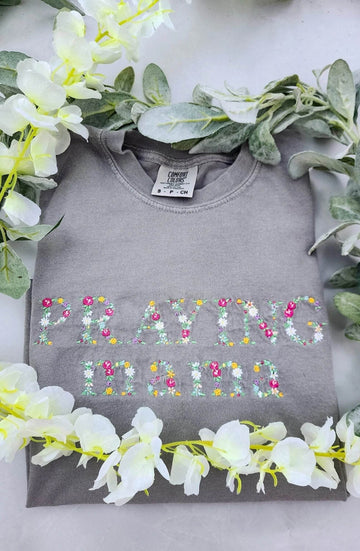 A long sleeve gray t-shirt with the text 'PRAYING MAMA' embroidered in colorful letters, displayed flat with a floral arrangement around it.praying-mama-shirt