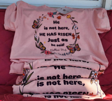 Stacked pink t-shirts with the text 'He is not here, HE HAS Risen' printed on them, placed on a red surface.