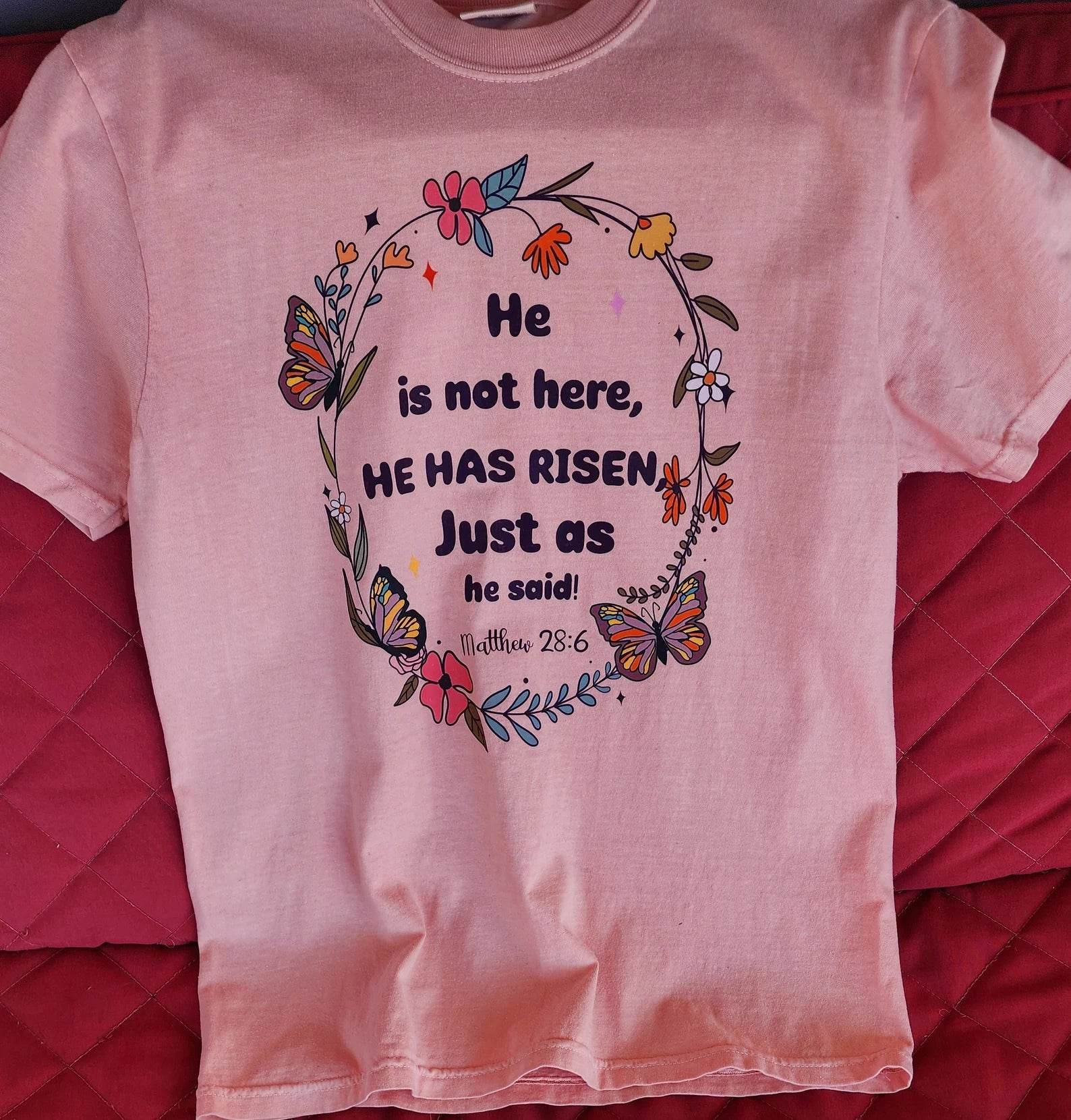 Stacked pink t-shirts with the text 'He is not here, HE HAS Risen' printed on them, placed on a red surface.