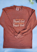 An inspirational t-shirt in brown color with the text 'Thank God, Trust God' printed on the front, displayed alongside denim jeans and black boots, set against a floral backdrop.