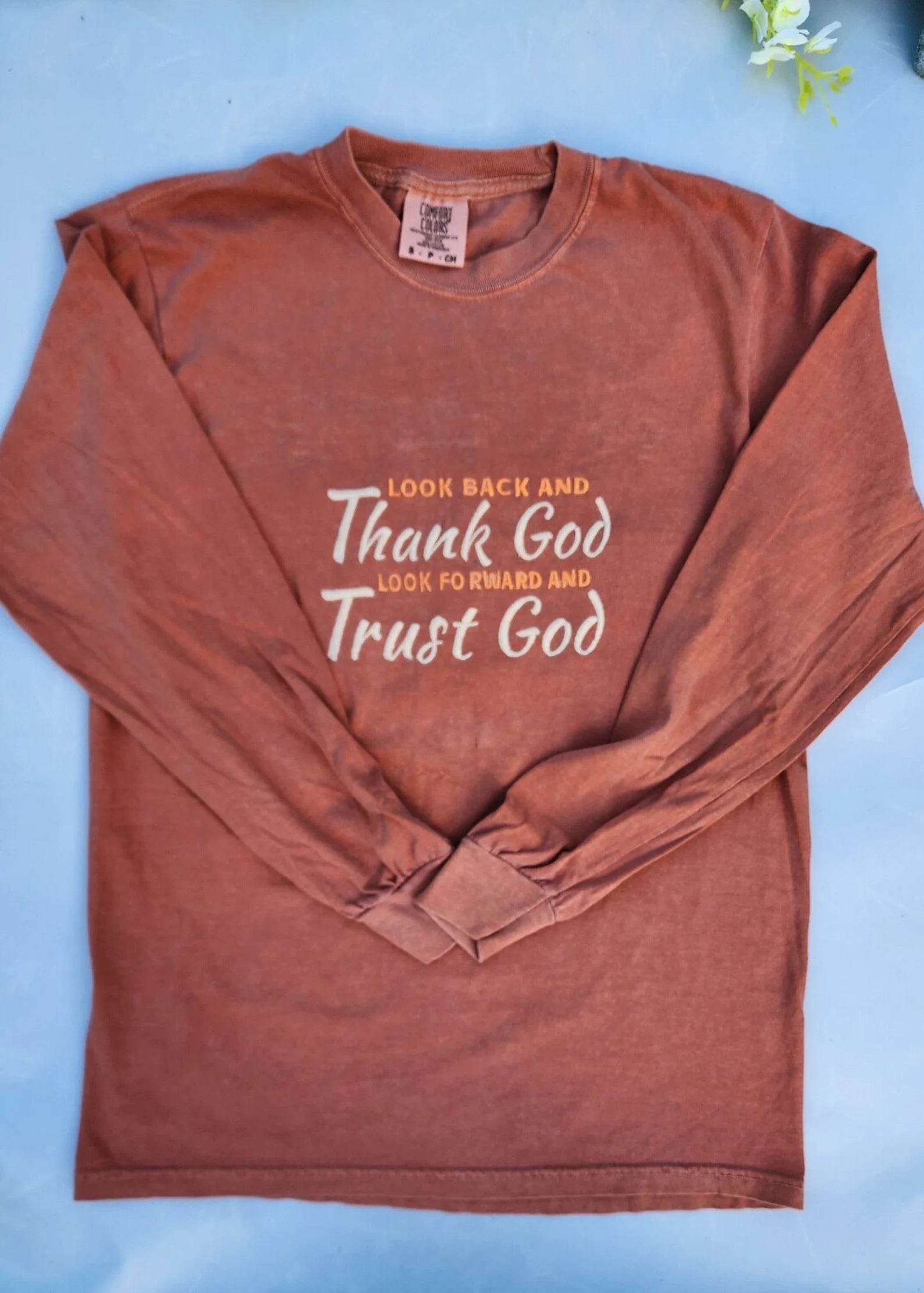 An inspirational t-shirt in brown color with the text 'Thank God, Trust God' printed on the front, displayed alongside denim jeans and black boots, set against a floral backdrop.
