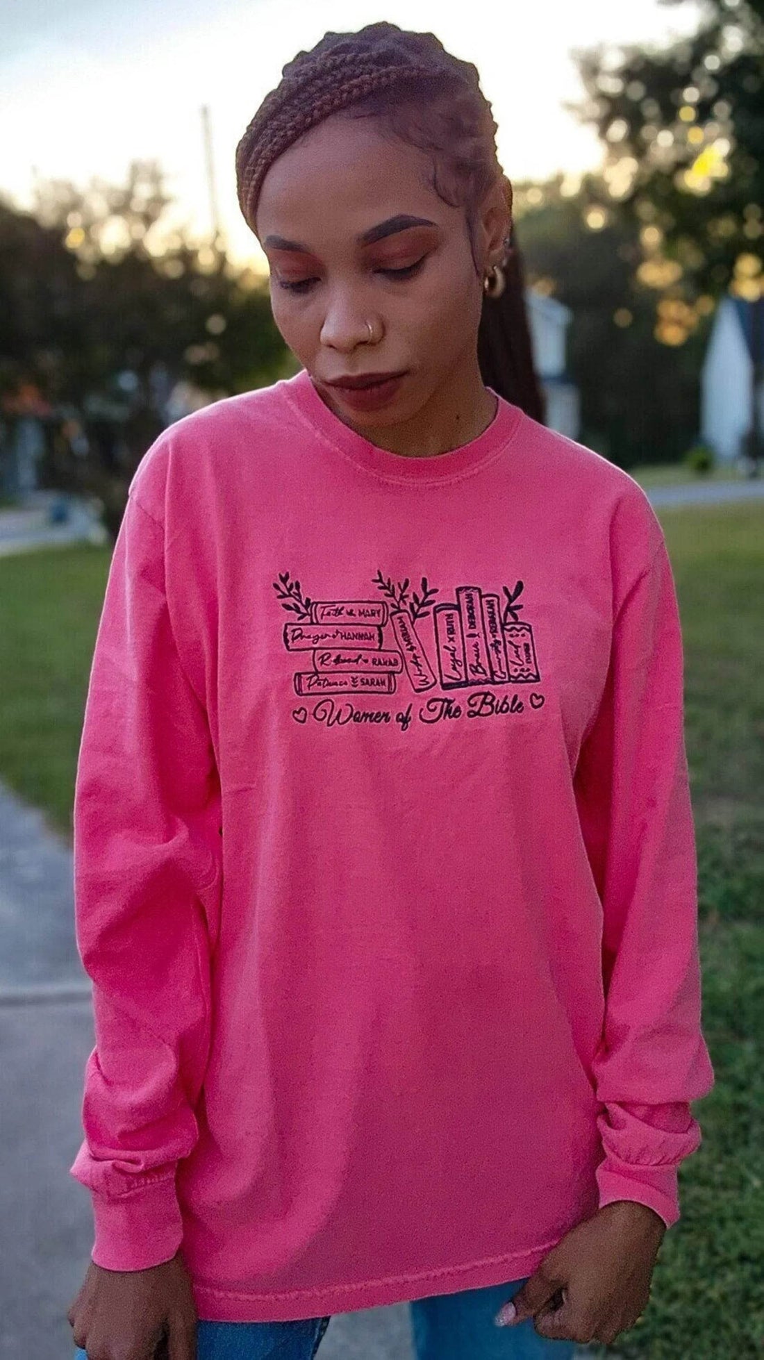 A woman wearing a pink long-sleeved t-shirt with embroidered text that reads 'Women of the Bible' and includes images of various women mentioned in the Bible.women-of-the-bible-shirt