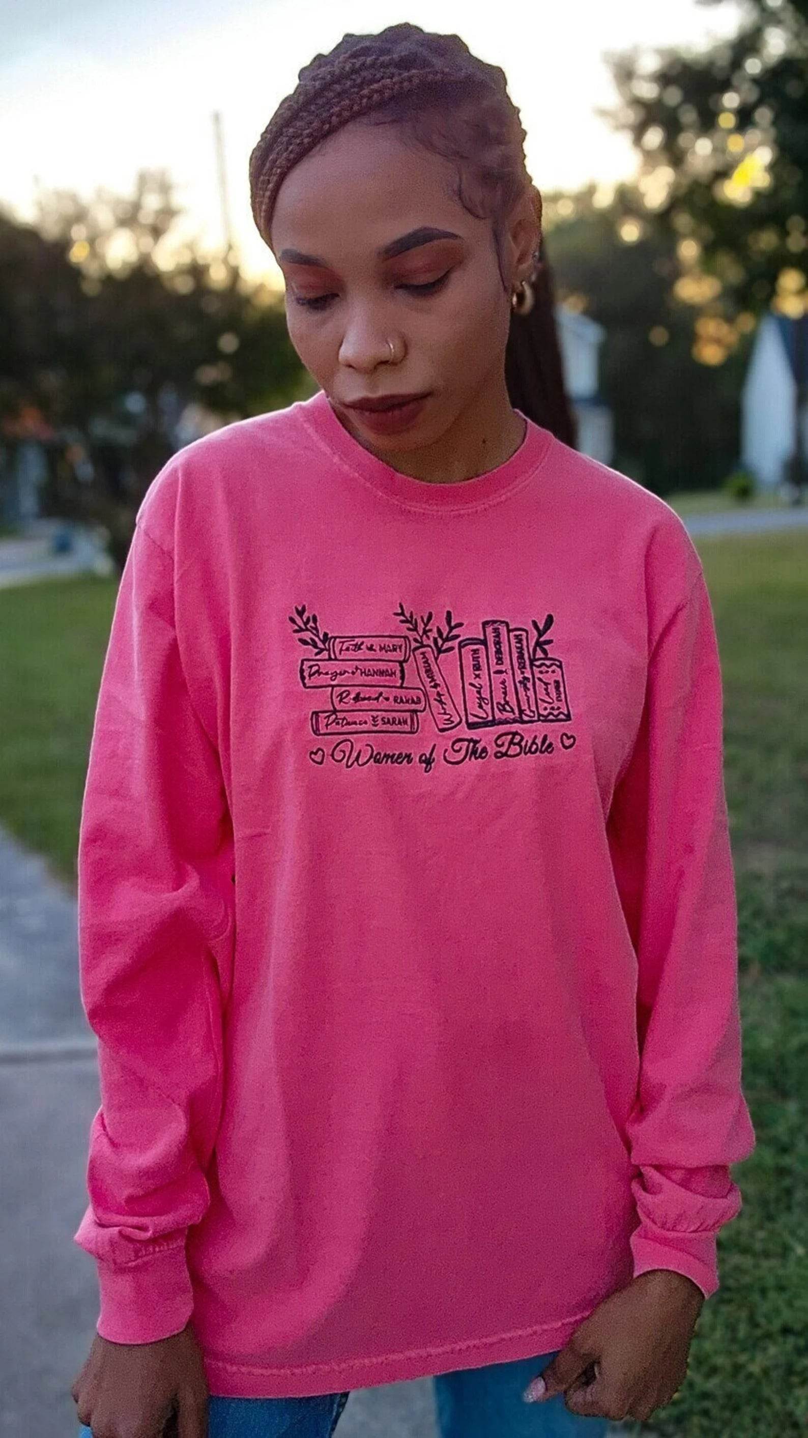 A woman wearing a pink long-sleeved t-shirt with embroidered text that reads 'Women of the Bible' and includes images of various women mentioned in the Bible.women-of-the-bible-shirt