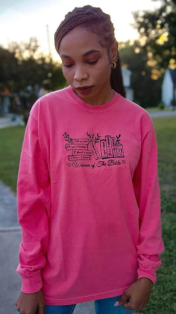A woman wearing a pink long-sleeved t-shirt with embroidered text that reads 'Women of the Bible' and includes images of various women mentioned in the Bible.women-of-the-bible-shirt