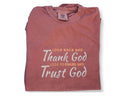 An inspirational t-shirt in brown color with the text 'Thank God, Trust God' printed on the front, displayed alongside denim jeans and black boots, set against a floral backdrop.