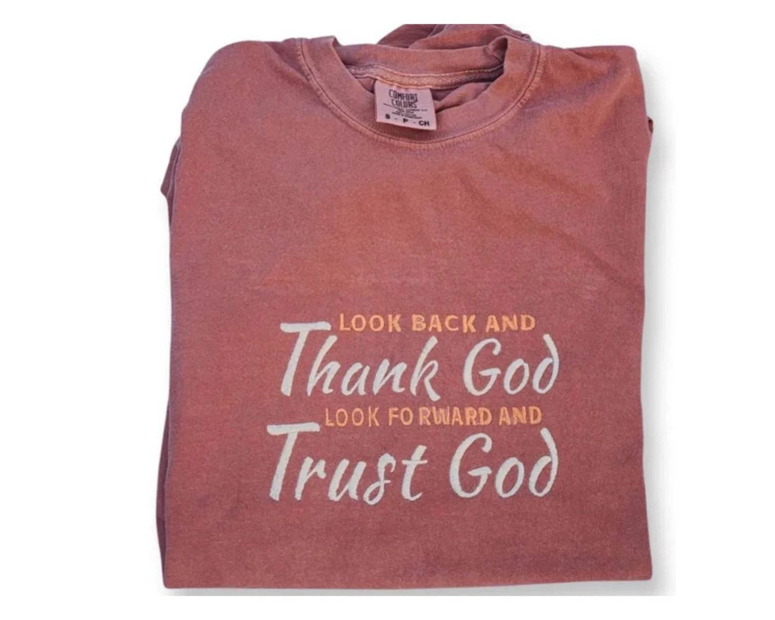 An inspirational t-shirt in brown color with the text 'Thank God, Trust God' printed on the front, displayed alongside denim jeans and black boots, set against a floral backdrop.