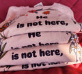 Stacked pink t-shirts with the text 'He is not here, HE HAS Risen' printed on them, placed on a red surface.