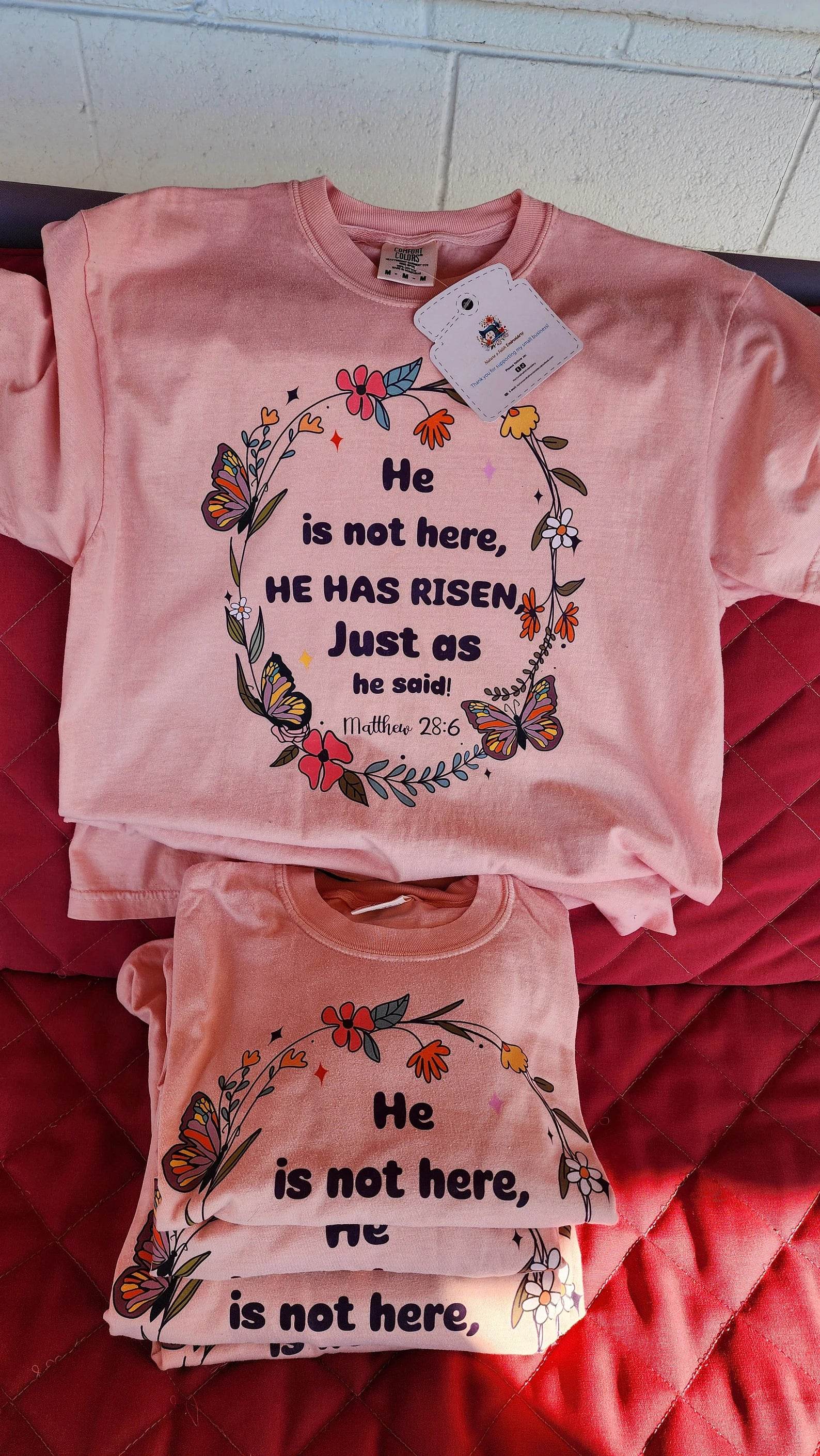 Stacked pink t-shirts with the text 'He is not here, HE HAS Risen' printed on them, placed on a red surface.