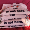 Stacked pink t-shirts with the text 'He is not here, HE HAS Risen' printed on them, placed on a red surface.