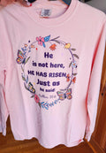Stacked pink t-shirts with the text 'He is not here, HE HAS Risen' printed on them, placed on a red surface.