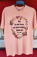 Stacked pink t-shirts with the text 'He is not here, HE HAS Risen' printed on them, placed on a red surface.