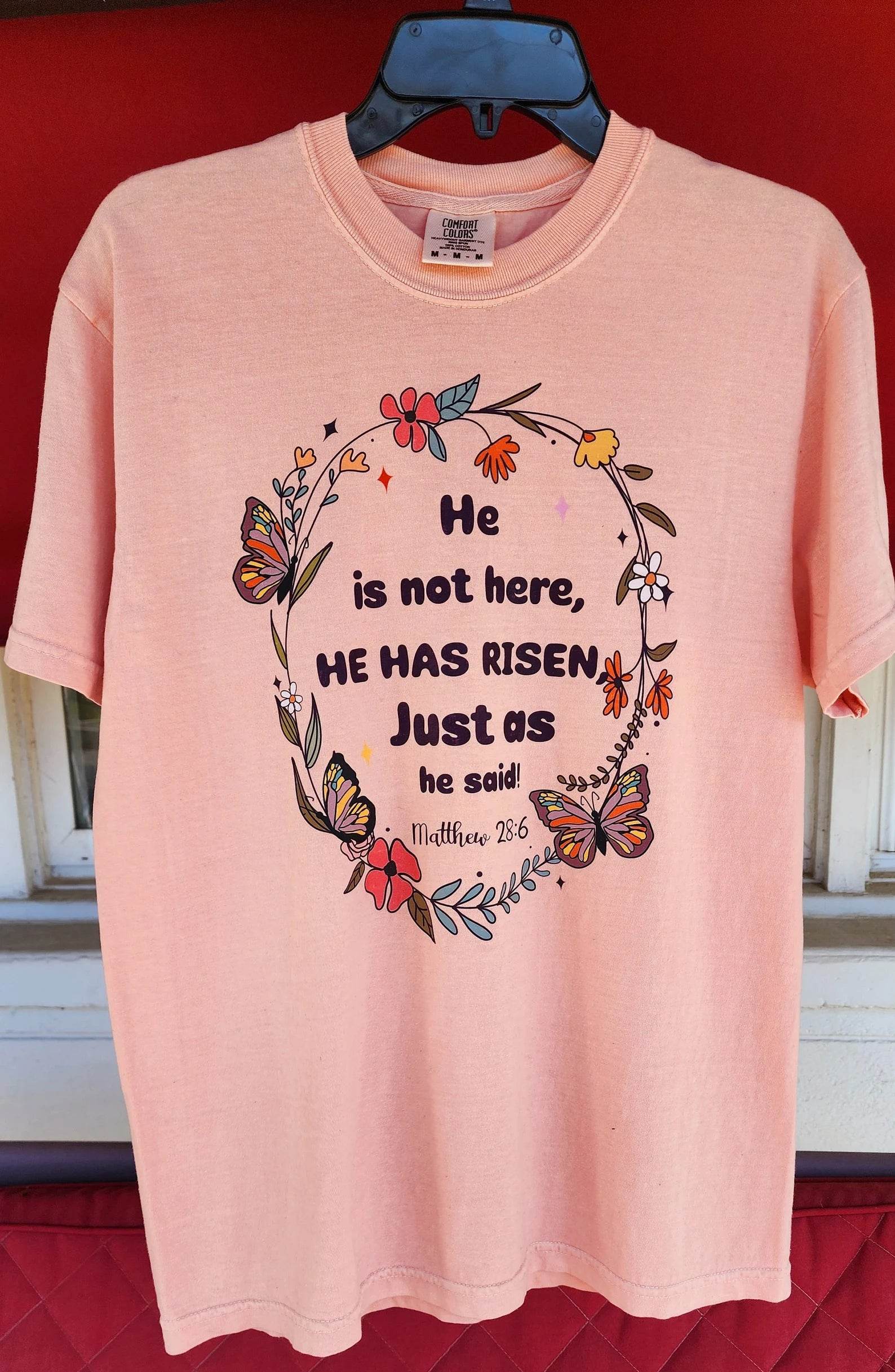 Stacked pink t-shirts with the text 'He is not here, HE HAS Risen' printed on them, placed on a red surface.