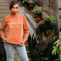 An inspirational t-shirt in brown color with the text 'Thank God, Trust God' printed on the front, displayed alongside denim jeans and black boots, set against a floral backdrop.