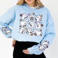 A woman wearing a light blue sweatshirt with a spooky nurse-themed design, including illustrations of ghosts and medical equipment, paired with black pants.