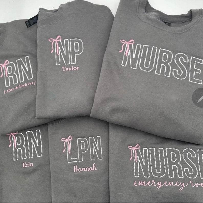A collection of custom embroidered grey nurse shirts with various titles and names embroidered on the left side of the chest.