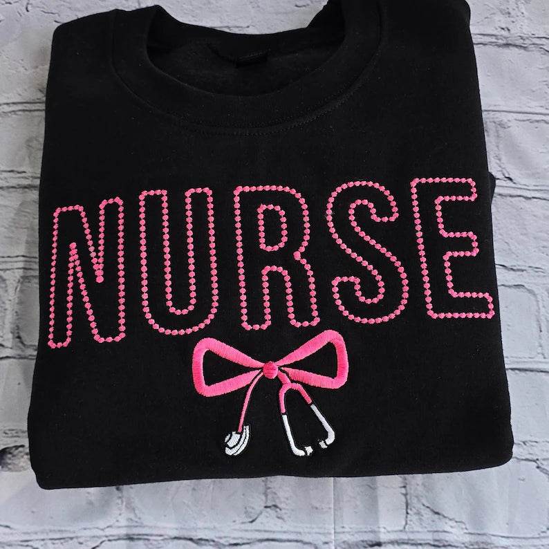 Black sweatshirt with embroidered pink text spelling 'NURSE' and a heart-shaped bow design underneath.
